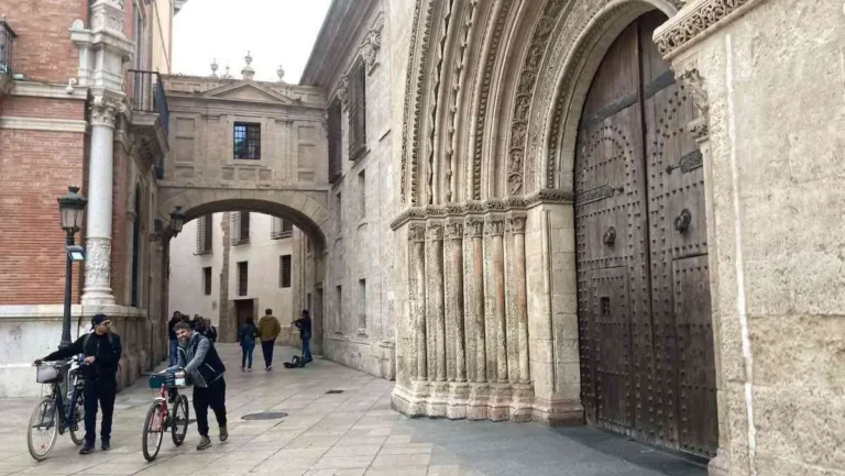 Two cyclists walk their bikes past the arched Cathedral doorway at Plaza Arzobispo in Valencia Spain