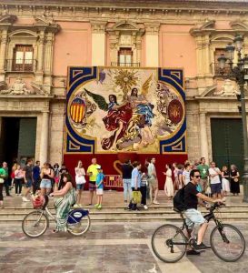 Cyclists and pedestrians in front of a large Corpus Christi decoration in front of Valencia's Basilica
