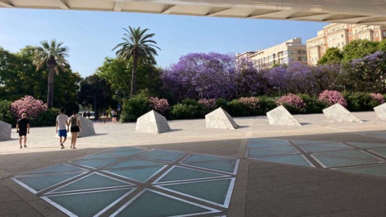 Pedestrians walk under the Alameda Bridge towards Turia Park trees with pink flowers