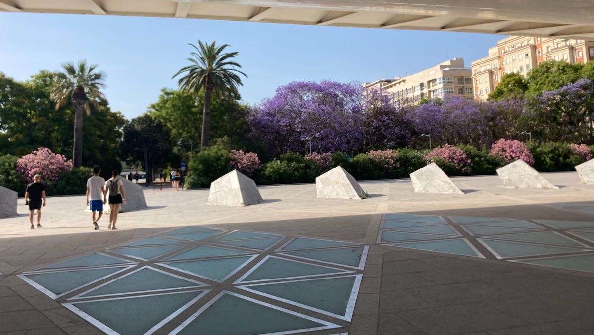 Pedestrians walk under the Alameda Bridge towards Turia Park trees with pink flowers