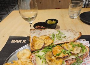 Open baguette sandwich served on a plate with a glass of white wine and small bowl of picles