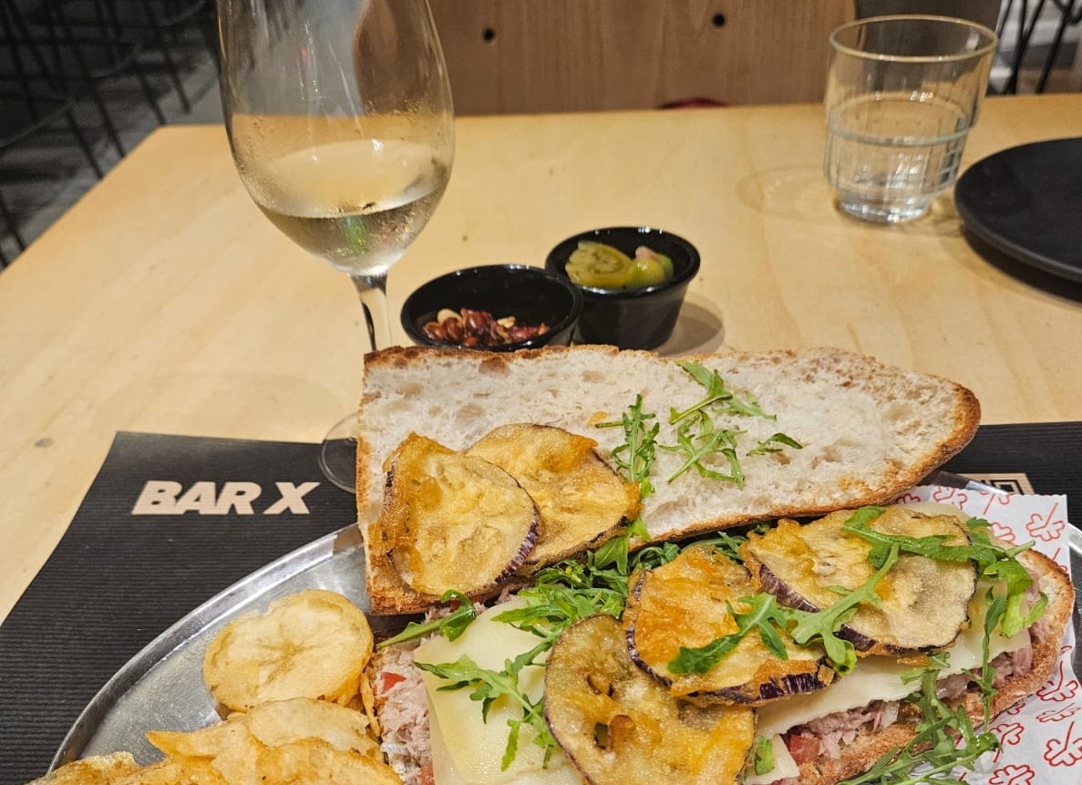 Open baguette sandwich served on a plate with a glass of white wine and small bowl of picles