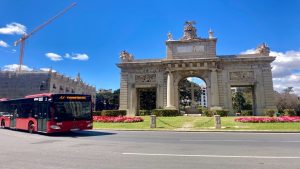 A red bus circling a roundabout with an arched monument in downtown Valencia, Spain