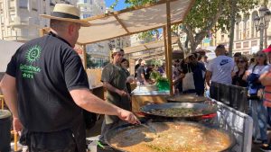 A man with black shirt and straw hat stirs a large pan of paella at a festival for Valencian rice