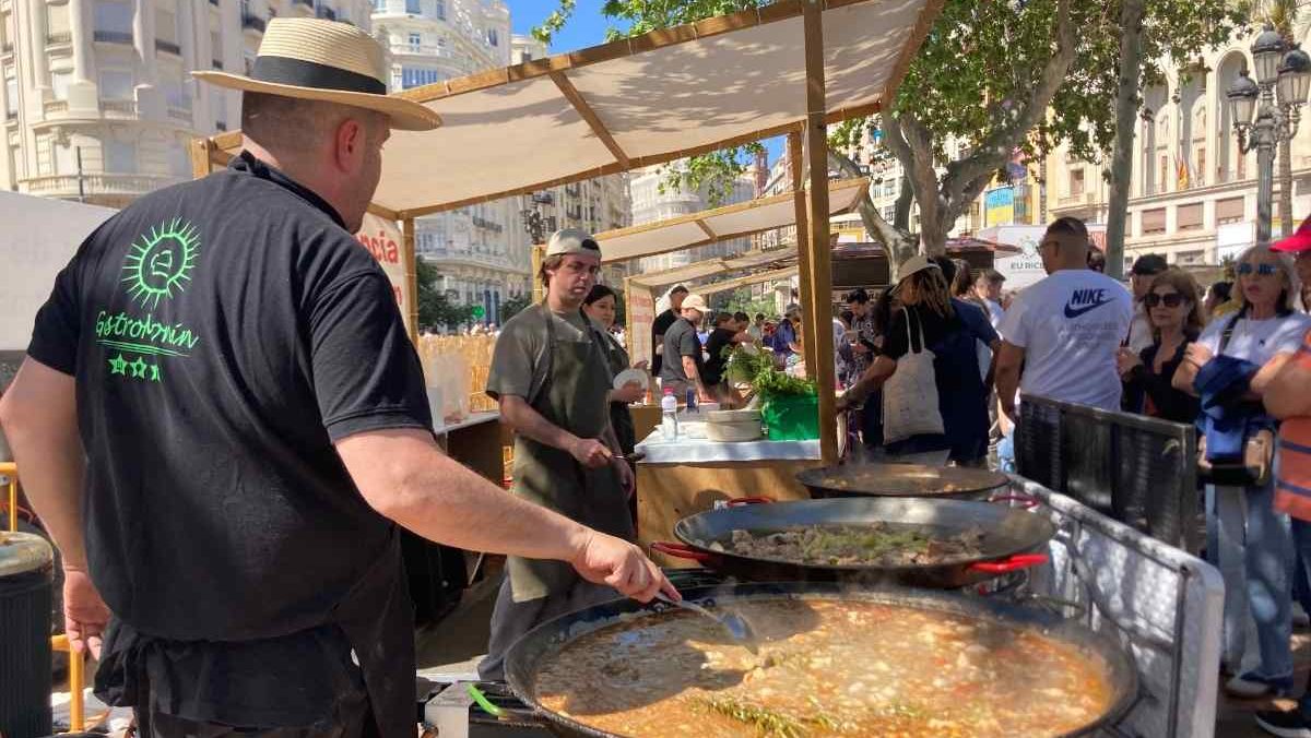 A man with black shirt and straw hat stirs a large pan of paella at a festival for Valencian rice
