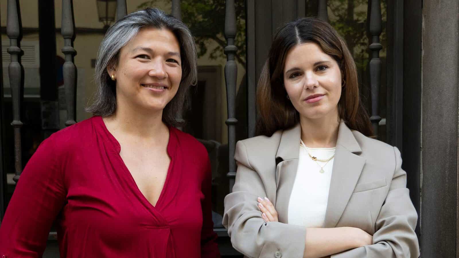 Atika Shubert, on the left in a red top, and Mireille Morejon, with dark hair in a beige blazer over a white top, creators of Pont de Valencia, standing in front of a wrought-iron gate.