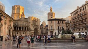 Pedestrians walk through Valencia's Plaza Virgen at sunset with a fountain and the back of the city's Cathedral visible.