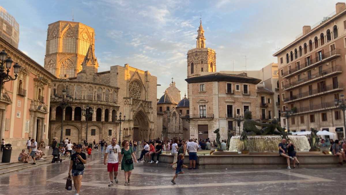Pedestrians walk through Valencia's Plaza Virgen at sunset with a fountain and the back of the city's Cathedral visible.