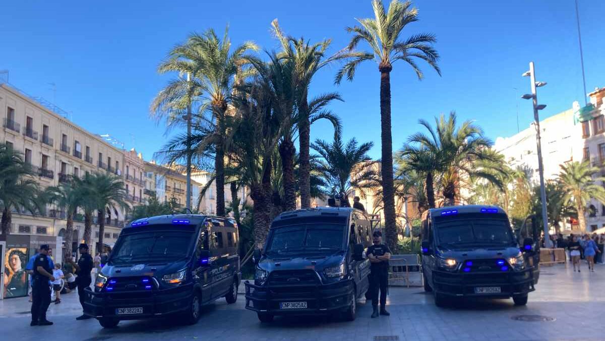 Three anti-riot police vans stand by under the palm trees of Plaza de la Reina on a sunny day in October 2026.