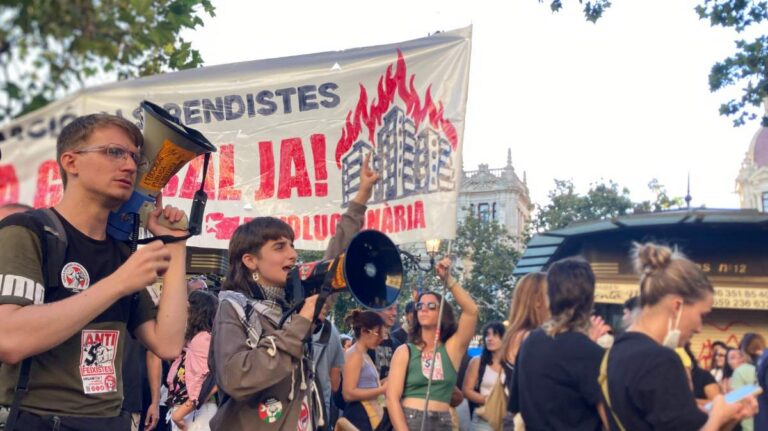 Protesters carrying loudspeakers and banners rally in front of a camp for housing rights in front of Valencia's Plaza del Ayuntamiento.